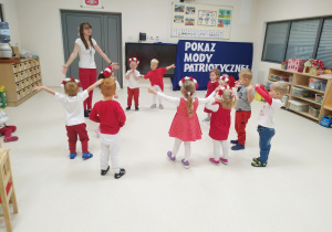 Dzieci i pani ubrani na biało - czerwono tańczą w kole z rozłożonymi rękami.